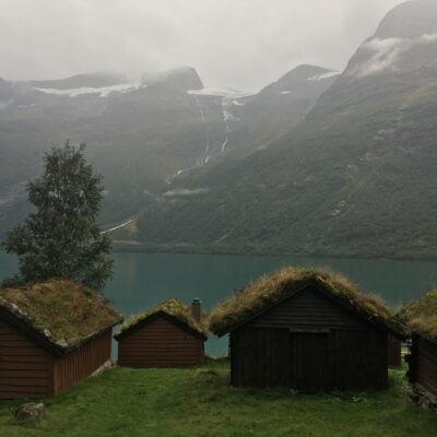 spowity mgłą krajobraz, na pierwszym planie drewniane domki z dachami pokrytymi trawą, w tle jezioro i wznoszące się nad nim szczyty