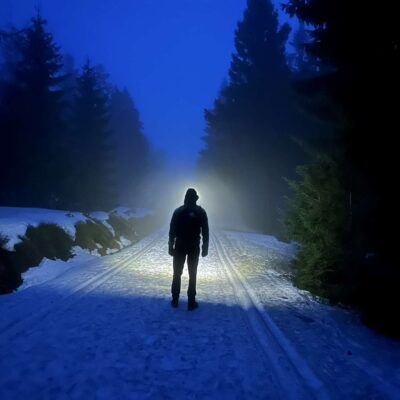 ciemno, zaśnieżony szlak wśród drzew, na środku drogi stoi mężczyzna oświetlający drogę czołówką