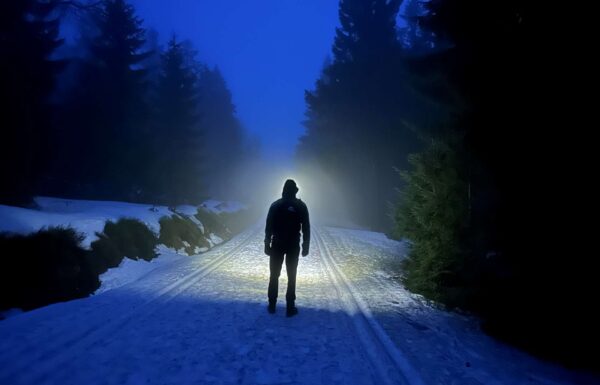 ciemno, zaśnieżony szlak wśród drzew, na środku drogi stoi mężczyzna oświetlający drogę czołówką