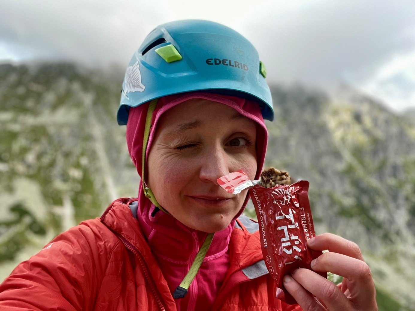 kobieta w niebieskim kasku wspinaczkowym, na tle Tatr Wysokich, spożywa baton This-1 i puszza oko do czytelnika