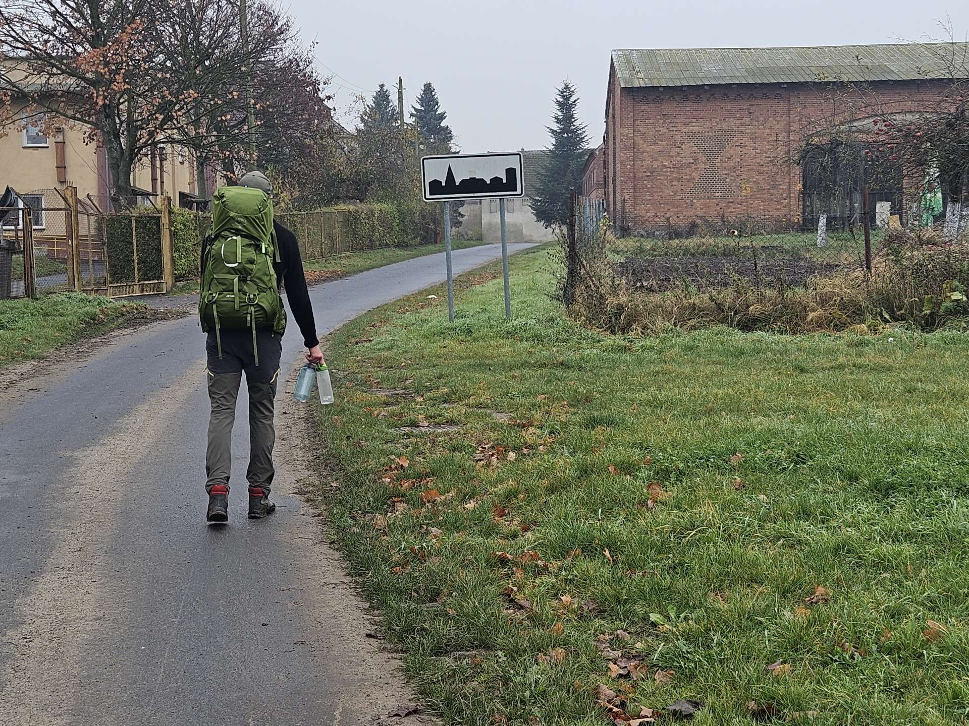 widziany z tyłu podróżnik z plecakiem i butelkami w dłoni, wśród zabudowań